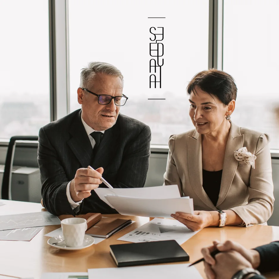 Two business professionals discussing documents at a conference table in a modern office setting, with a city view in the background.