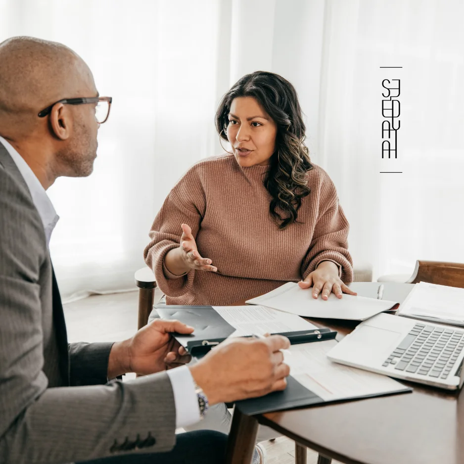 Woman in a brown sweater engaged in discussion with a man in a suit at a table, with documents and a laptop present.