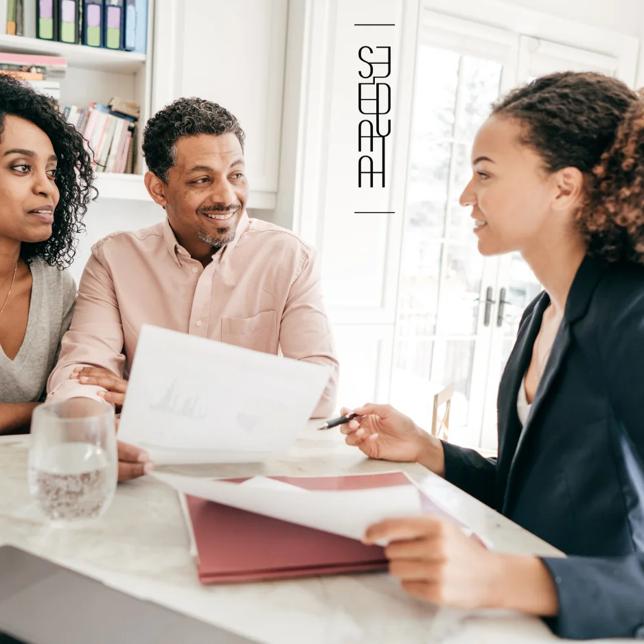 A group of three people engaged in a discussion over documents and charts at a table, with a modern and bright interior setting.
